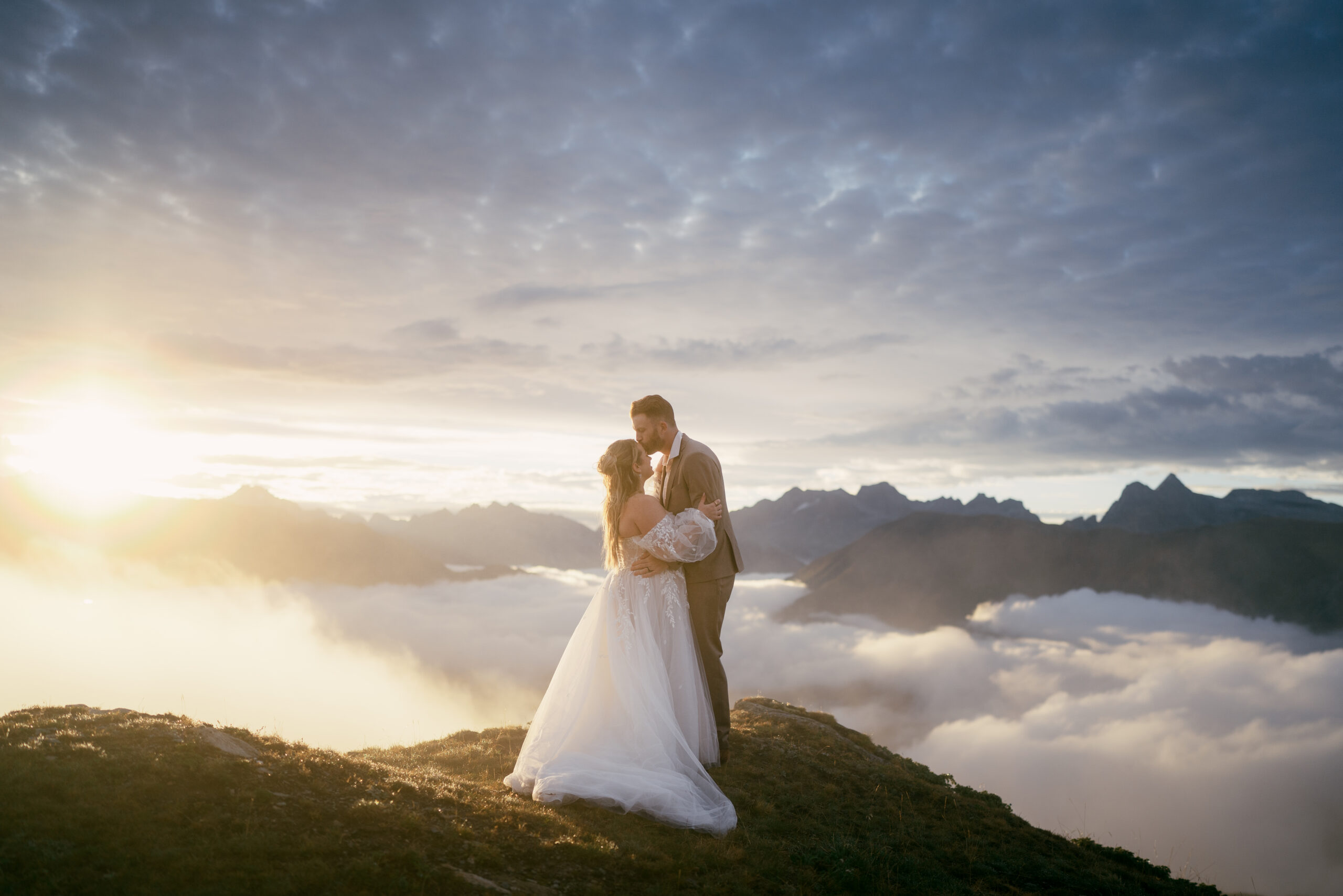 a bride and groom kiss on a mountain at sunrise, warm light touching the peaks around them. They are standing above the clouds, which have remained low in the valley.