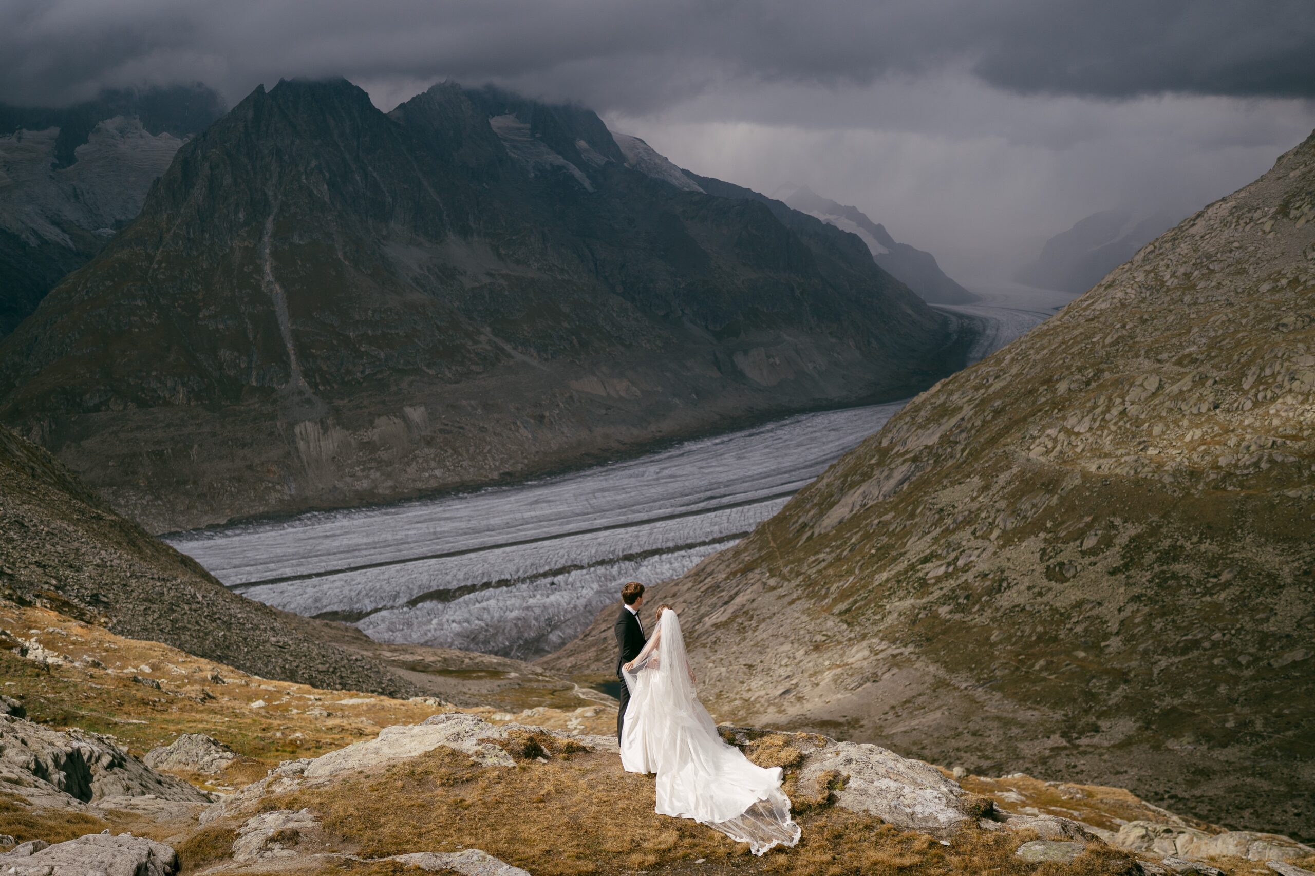 A bride and groom stand on a rock overlooking a glacier as moody dark clouds enshroud the mountains.