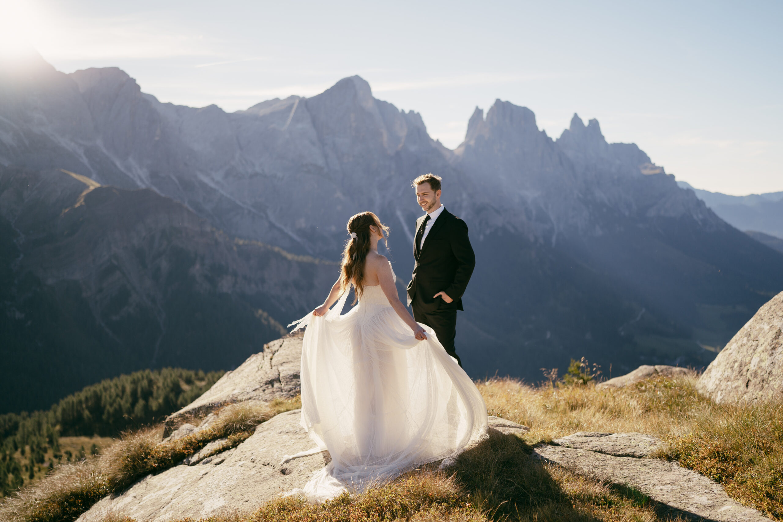 A bride twirls her dress while her husband smiles at her as they stand on a mountain. Warm sunshine spills over the jagged peaks behind them.